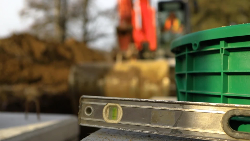 Closeup footage in shallow focus as a well used builder spirit level is seen resting on a septic tanks, a blurry excavator digs a trench in background Royalty-Free Stock Footage #1047304240