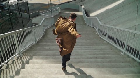 Happy Young Man Actively Dancing While Walking down the stairs. He's Wearing a brown long coat. Scene Shot in an Urban Park Next to Business Center. Slow motion. Shooting from the air.