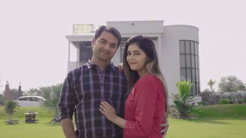 A happy and smiling couple holding each other standing in front of their dream home. A married husband/wife outside the newly constructed bungalow looking at the camera with a smile. 