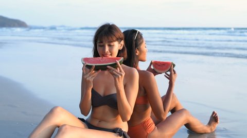 Holiday concept. Two girls in bikinis eating watermelon on the beach. 4k Resolution.