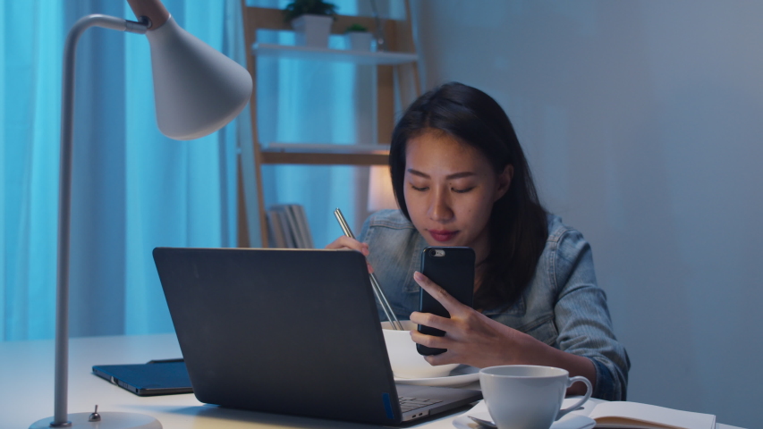 Freelance smart business women eating instant noodles live on social media via mobile phone in living room at home at night. Happy young Asian girl sitting on desk work overtime, enjoy relax time. | Shutterstock HD Video #1047326110