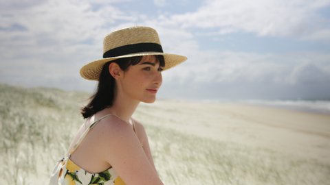 Beautiful brunette woman wearing a hat sitting on a beach looking out at the ocean in Australia with soft natural lighting. Medium shot on 4k RED camera.
