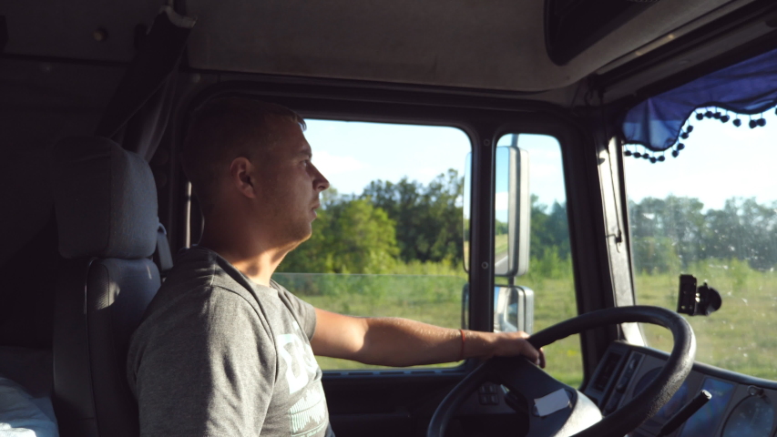 Man holds hand on the steering wheel and driving truck through countryside. Truck driver profile. Trucker sitting at the wheel of the car. Caucasian guy looks carefully at the road. Side view Close up