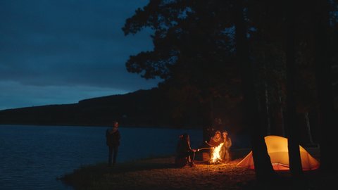 group of friends sitting by campfire roasting marshmallows camping in forest by lake an night chatting sharing warmth enjoying outdoor adventure 4k
