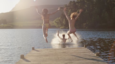 happy friends running jumping off jetty in lake at sunset having fun splashing in water enjoying freedom sharing summertime adventure