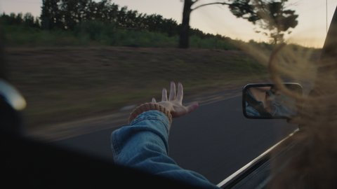 young woman in car holding hand out window feeling wind blowing through fingers driving in countryside on road trip travelling for summer vacation enjoying freedom on the road at sunset