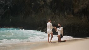 Young handsome man standing on one knee and doing proposal to his beautiful girlfriend on tropical paradise beach. Touching moment of couple getting engaged