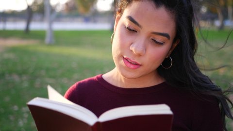 Close up on a young hispanic woman college student reading the pages of a red story book or novel outdoors SLOW MOTION.