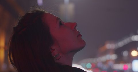 Profile portrait of Caucasian girl looking up outdoors shallow city traffic lights background copy space. Side view of young woman gazing to skies at night slow motion text. Freedom happiness dreams