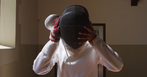 Front view of a confident focused mixed race female fencer athlete during a fencing training in a gym, wearing jacket and plastron, preparing for a fencing duel, putting mask on