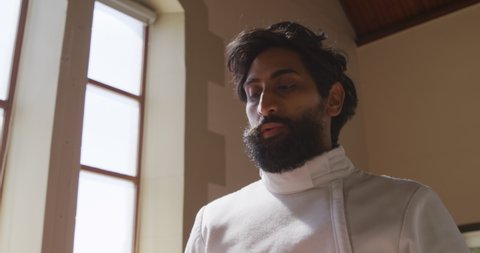 Side view close up of a confident focused mixed race male fencer athlete during a fencing training in a gym, wearing jacket and plastron, preparing for a fencing duel, putting mask on, holding an epee