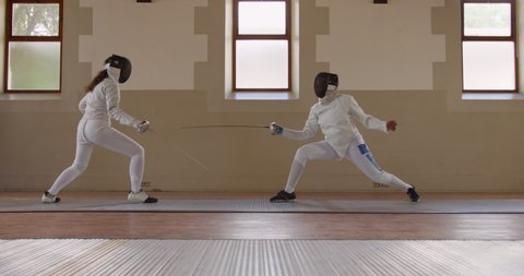Side view of a Caucasian and a mixed race female fencer athletes during a fencing training in a gym, sparring in fencing duel wearing masks, jumping taking aim at each other with their epees in slow