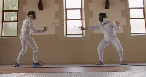 Side view of a Caucasian and a mixed race male fencer athletes during a fencing training in a gym, sparring in fencing duel wearing masks, jumping taking aim at each other with their epees, one man