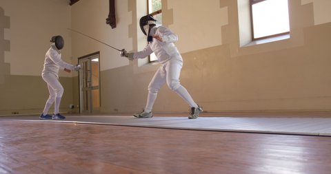 Low angle side view of a Caucasian and a mixed race male fencer athletes during a fencing training in a gym, sparring in fencing duel wearing masks, jumping taking aim at each other with their epees