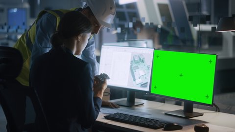 Supervisor Talks to Female Engineer Who's Working on a Computer, Two Monitor Screens Show Chroma Key, Green Screen Display and Processor Chip Design. Industry 4.0 Modern Electronics Production Factory