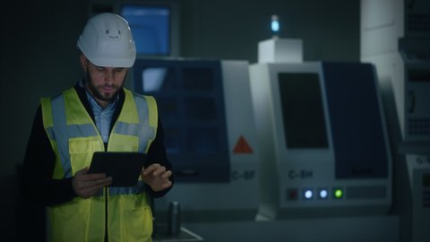 Professional Engineer Wearing Safety Vest and Hardhat Walks Through Modern Factory Workshop, Uses Digital Tablet Computer. Handsome Professional Working in Industrial Facility with High Tech Machinery