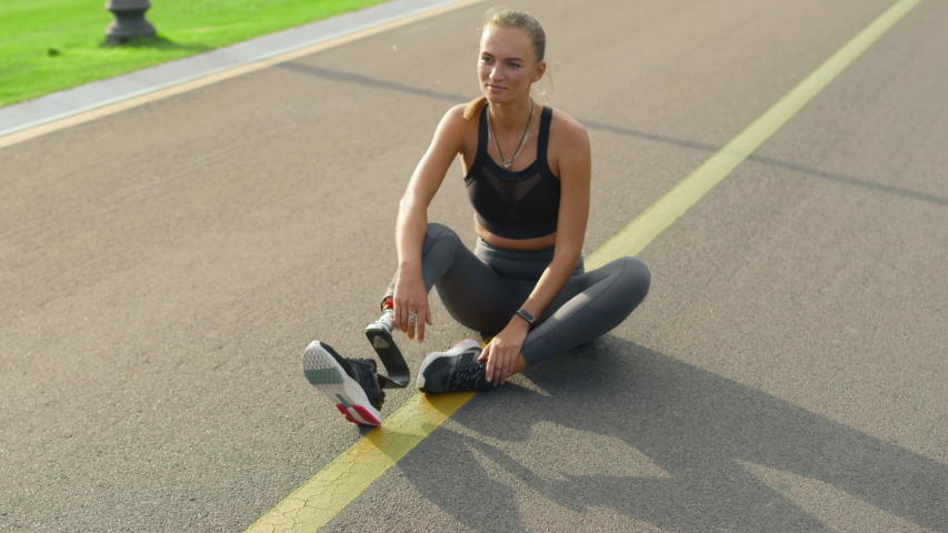Smiling woman with artificial limb sitting on road. Cheerful girl relaxing after training in park. Fit lady in sportswear taking break after workout outdoors in slow motion