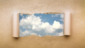 Creative 4k time laps video of fast moving clouds in the blue sky that are visible through a hole with torn edges in old retro grunge vintage paper.