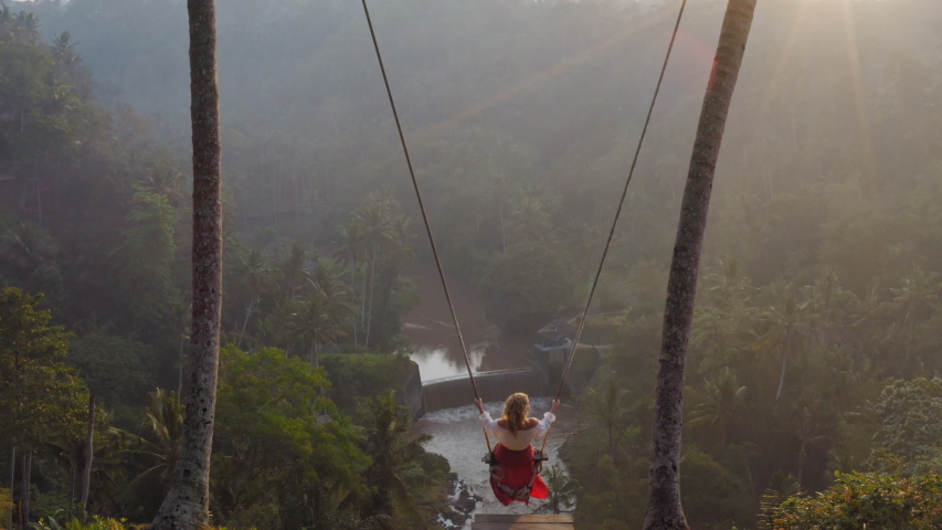 aerial view woman swinging over tropical rainforest at sunrise sitting on swing with view of river enjoying having fun on holiday travel freedom