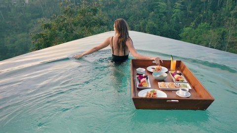travel woman having breakfast in swimming pool enjoying exotic food at luxury hotel spa with view of tropical jungle at sunrise