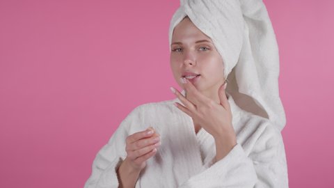 Portrait shot of beautiful young woman in shower robe and towel standing against pink background and applying lip scrub while looking at camera