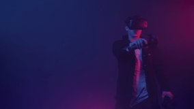 Attractive Man in Virtual Reality Glasses Moving Hand with Controller Joystick in it, Standing in the Room with Neon Lighting Colors. Futurictic Background.