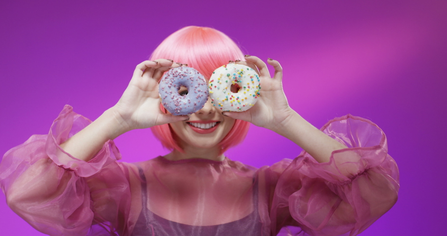 Close up of pretty Caucasian young woman in stylish outfit closing eyes with two donuts and smiling joyfully. Portrait of beautiful happy girl playing with sweets on velvet wall background.
