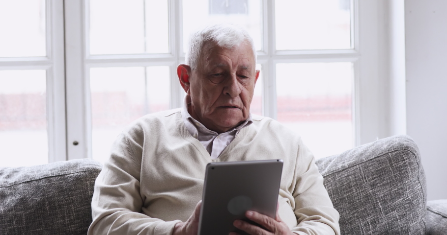 Smiling senior elder 80s man using digital tablet computer pad modern tech relax on sofa. Happy retired old grandfather browse internet read e book in app chatting online with distance doctor at home. Royalty-Free Stock Footage #1047422629