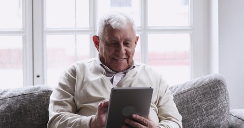 Smiling senior elder 80s man using digital tablet computer pad modern tech relax on sofa. Happy retired old grandfather browse internet read e book in app chatting online with distance doctor at home.