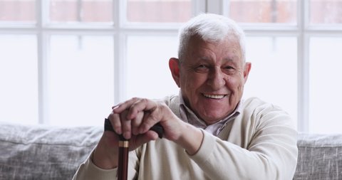Happy older senior adult man hold cane sit on sofa looking at camera. relaxed optimistic disabled retired elderly 80s grandfather dental smile posing with walking stick at nursing home, portrait