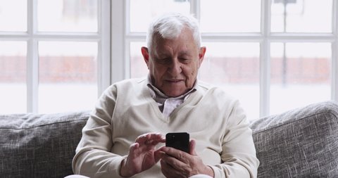 Smiling elder 80s adult grandfather learn mobile phone app sit on sofa, happy 70s senior man hold smartphone texting messages in social media reading news, old person using tech gadget concept at home