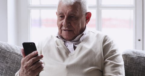 Happy senior elderly 70s man user holding smart phone watching mobile video calling online looking at screen relaxing on couch at home, older grandparent learn using modern technology gadget concept.