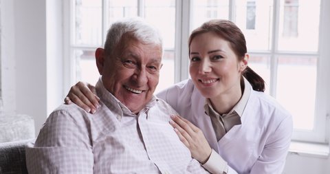 Caring smiling female caregiver embracing happy senior patient looking at camera. Young carer or nurse helping old elder care grandpa at nursing home, geriatric medical support concept, closeup