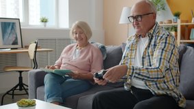 Woman is reading book while elderly man is playing video game at home on couch relaxing in free time. Retirement and leisure time activities concept.