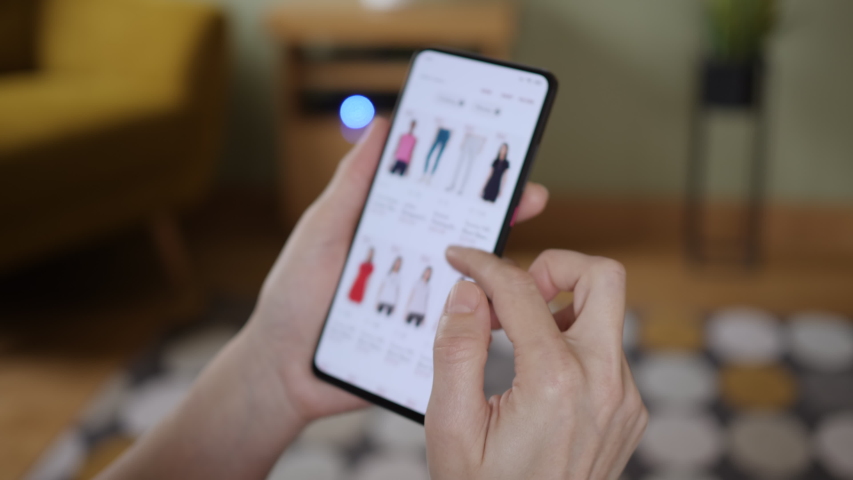 Woman Looks at Goods in Online Store Clothing. Buy Fashion Clothes Directly on Smartphone. Woman at Home Lying on Couch in Living Room Using Smartphone Buys in Internet Shop. | Shutterstock HD Video #1047443911