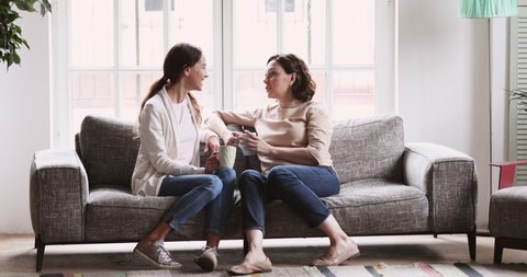Happy family middle aged old mother and young adult daughter chatting. Mum and grown child rest in cozy living room interior. Smiling two 2 age generations women friends talk drink tea on couch at