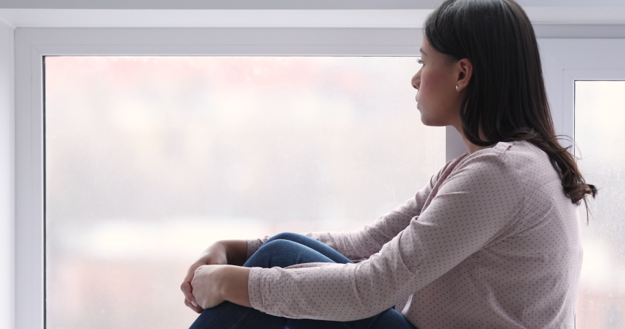 Thoughtful lonely millennial african woman sit on sill look through window. Sad sensitive mixed race lady introvert reflect think of solitude feel anxious in melancholic mood concept, closeup side | Shutterstock HD Video #1047458632