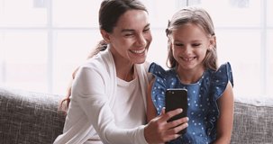Cheerful young adult mum show cute small kid daughter funny mobile video. Happy family mother and little preschool girl having fun use smartphone. Parent with child laugh looking at phone sit on sofa