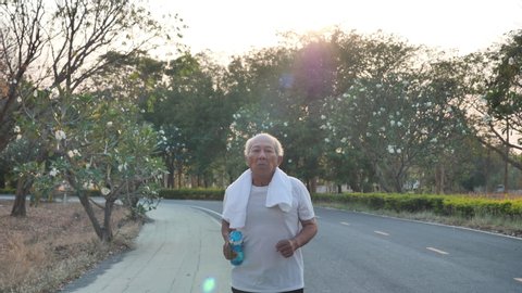 Front view Asian elderly exercise. Senior man running on road in public park that has sunshine in the evening. health lifestyle and exercise Concept. Slow Motion