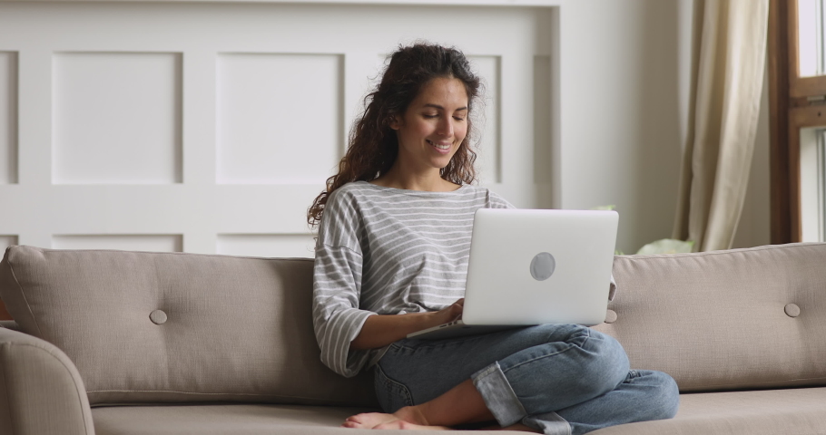 Pleasant young curly woman relaxing on sofa, using computer at home. Smiling millennial lady communicating with friends in social networks, chatting with boyfriend online, internet shopping on laptop. Royalty-Free Stock Footage #1047475810