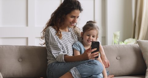 Smiling young mother cuddling adorable preschool daughter, making selfie photo together. Happy babysitter nanny using funny mobile applications, sitting on cozy couch with cute small child girl.