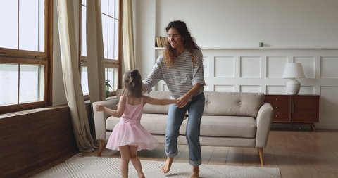 Full length overjoyed little child daughter wearing crown princess dress holding hands of smilig young nanny baby sitter mother, dancing twisting together on floor carpet in modern living room.