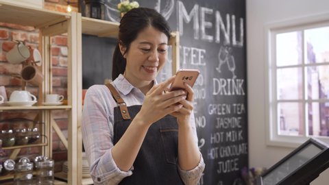 happy female coffee shop worker. young girl waitress in apron standing in counter bar and using cellphone chatting online laughing. asian woman barista relax texting message on mobile phone in cafe