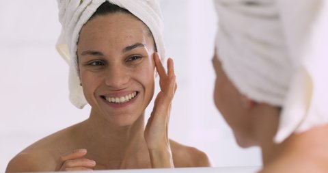 Head shot close up attractive happy young mixed race woman with towel on head looking at mirror touching skin, satisfied with its condition. Smiling millennial girl enjoying healthy moisturized face.