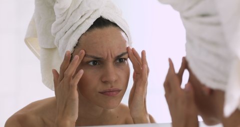 Frustrated young mixed race girl with towel on head looking at mirror, worrying about first wrinkles. Unhappy woman dissatisfied with skincare product effects or skin condition, feeling stressed.