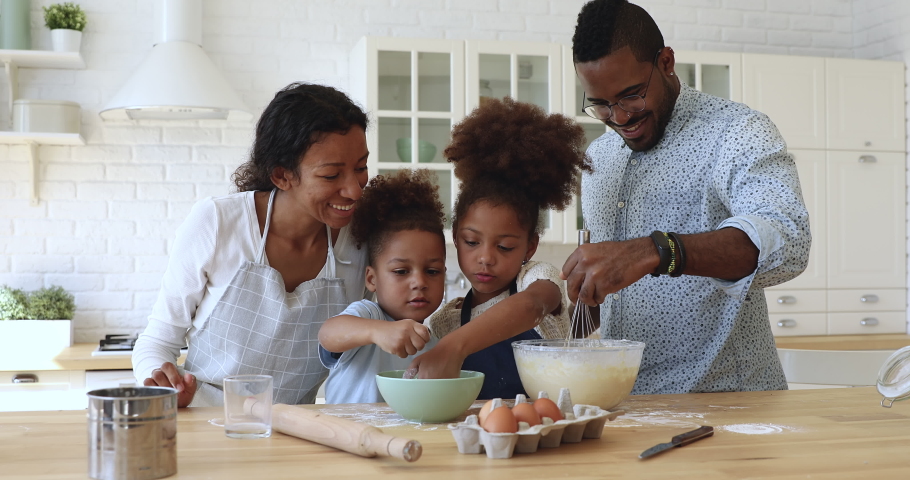 Happy full family having fun preparing pastry together at home. Smiling african dad whisking mixture in bowl while joyful little children adding flour, cooking homemade baking together at kitchen. Royalty-Free Stock Footage #1047485464