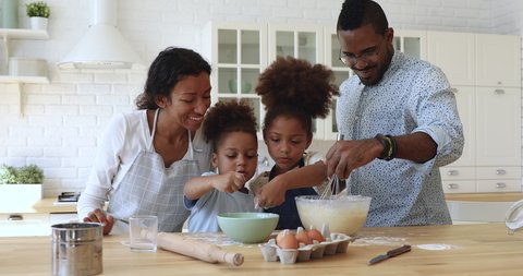 Happy full family having fun preparing pastry together at home. Smiling african dad whisking mixture in bowl while joyful little children adding flour, cooking homemade baking together at kitchen.