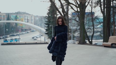 Attractive girl posing on camera in the city park. Beautiful model in black clothing walking outdoors. Lovely girl with opened fur coat. Slow motion.