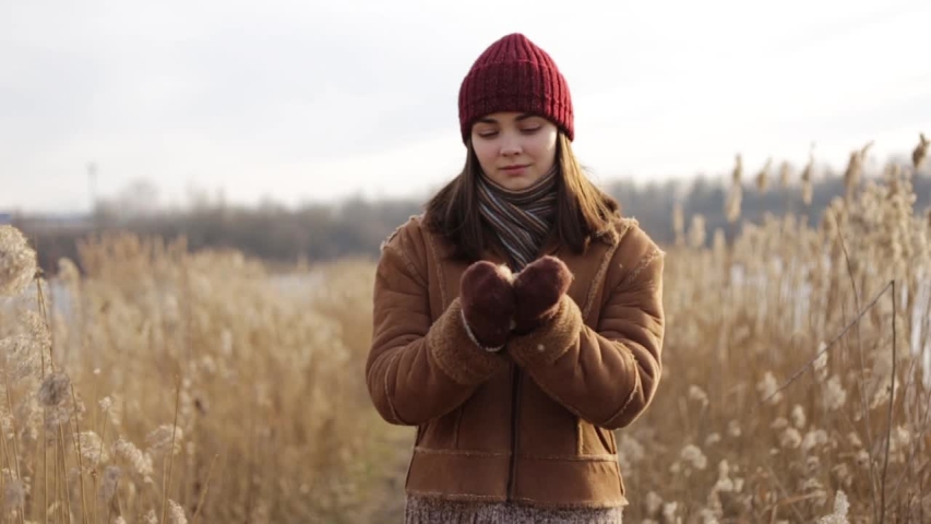 Young woman fluttering fluff in wind in field. Cheerful female standing in rural area in high grass and scattering cattail fluff in wind while laughing