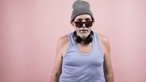 Hipster aged man in grey hat, blue tank top, sunglasses and headphones around neck. points with finger on his right biceps on pink background